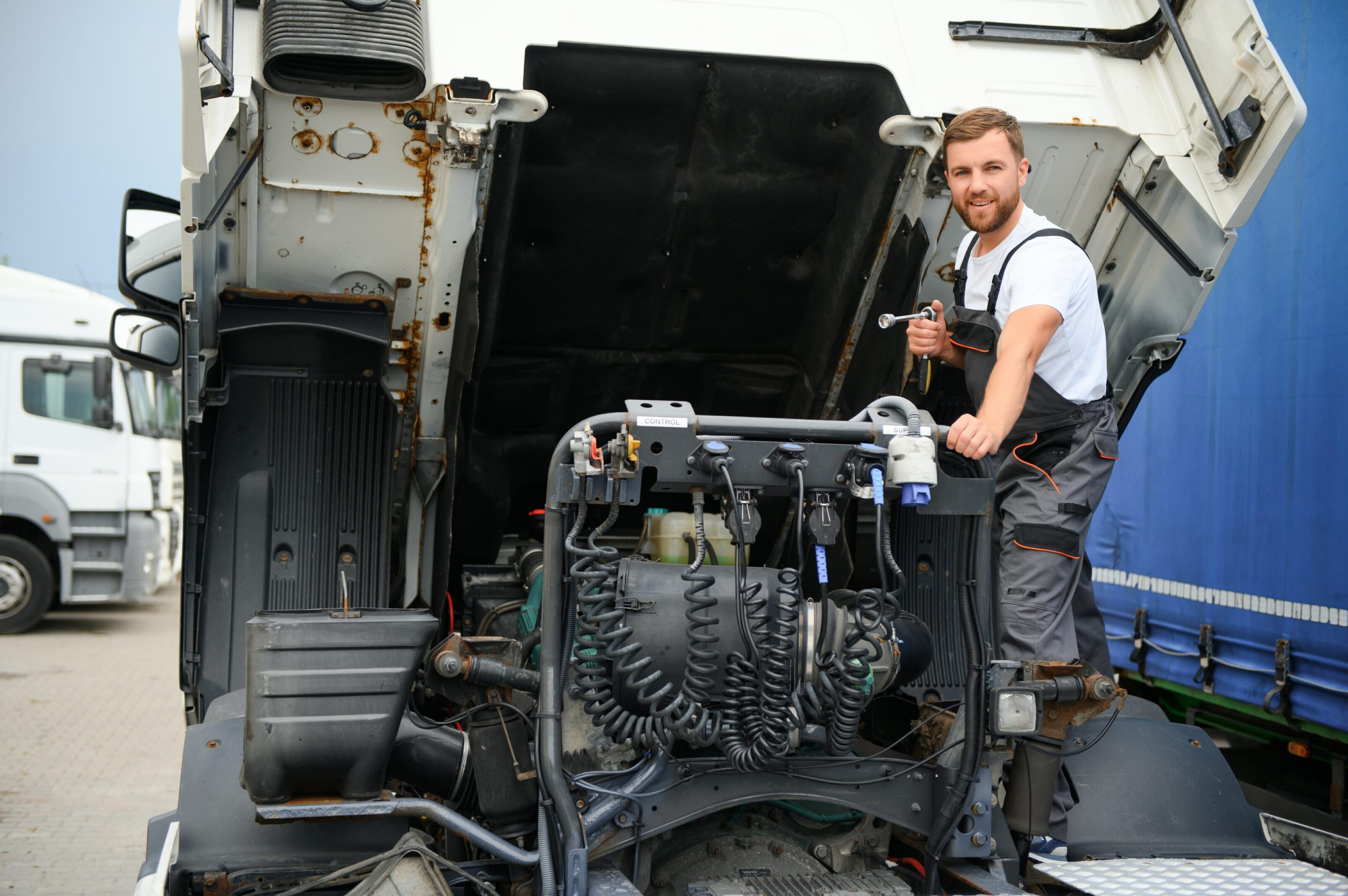 Mechanic working on the engine of a large truck with the cab tilted forward, holding a tool and looking at the camera.